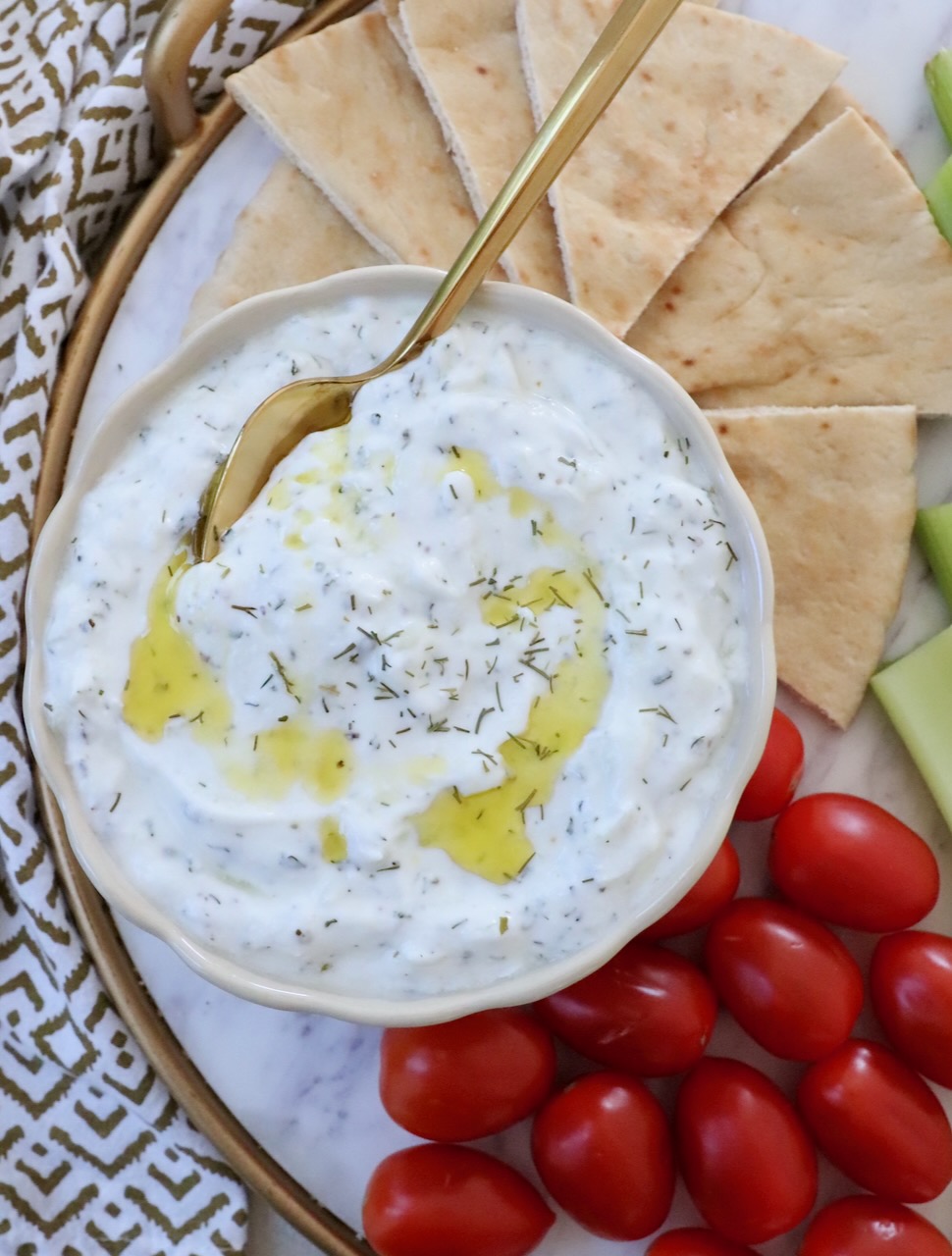 tzatziki sauce in a bowl with a spoon with olive oil drizzled on top and slices of pita bread and cherry tomatoes on the side