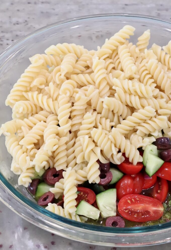 cooked rotini pasta, diced cucumber, tomatoes and olives in a large glass bowl