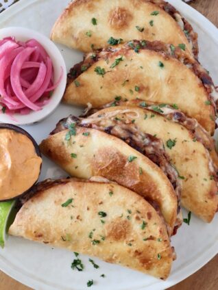 baked tacos stacked up on a plate with a side bowl of pickled onions and a bowl of chipotle sauce