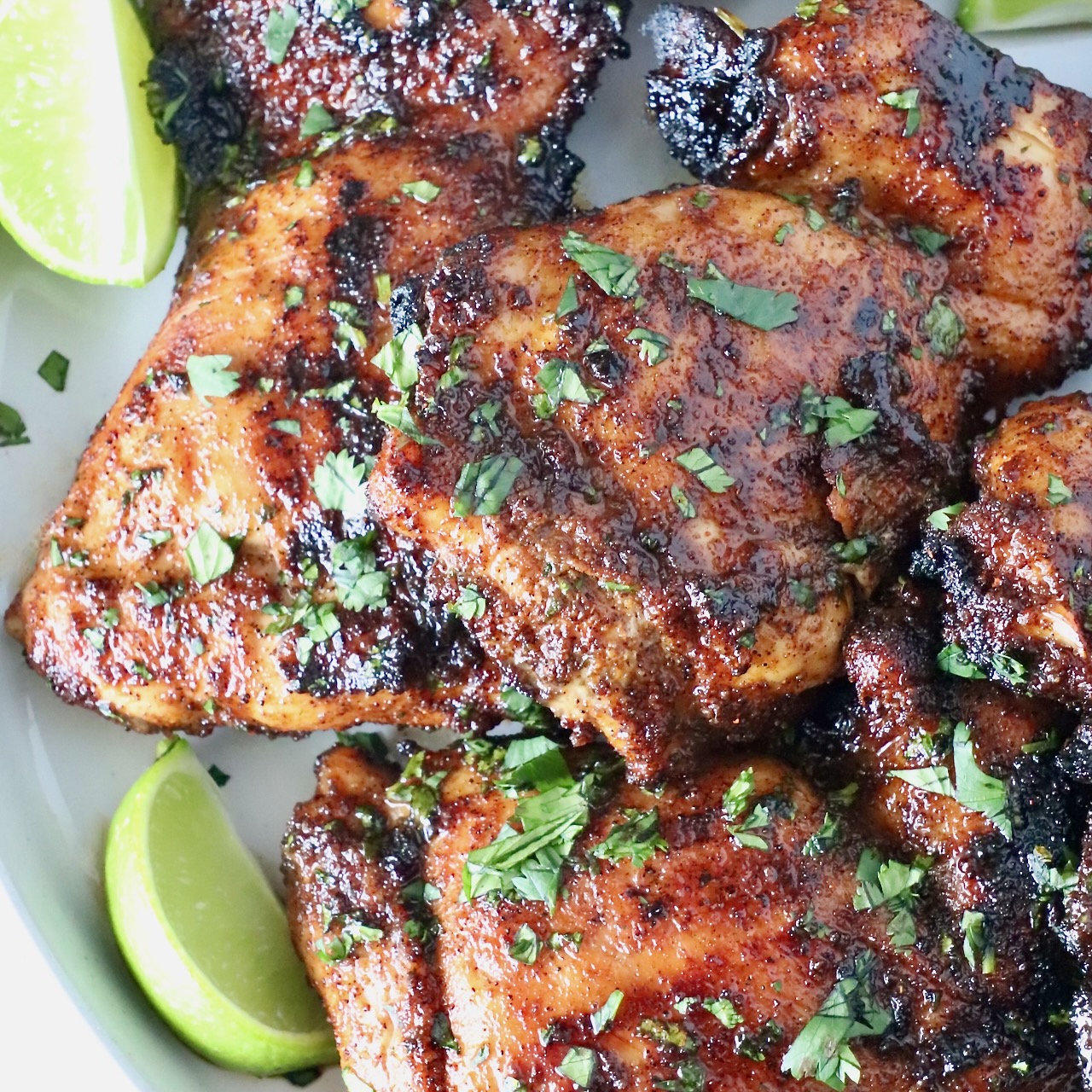 grilled chicken thighs stacked up on a plate with fresh cilantro and lime wedges