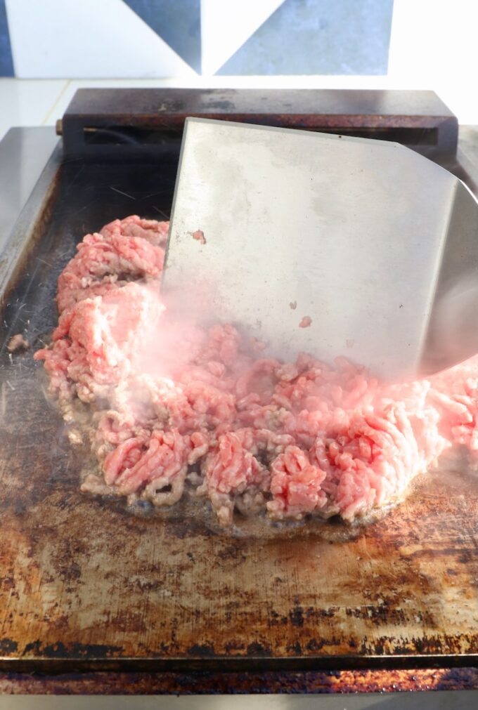 ground beef on a flat top griddle with a large metal spatula in the beef