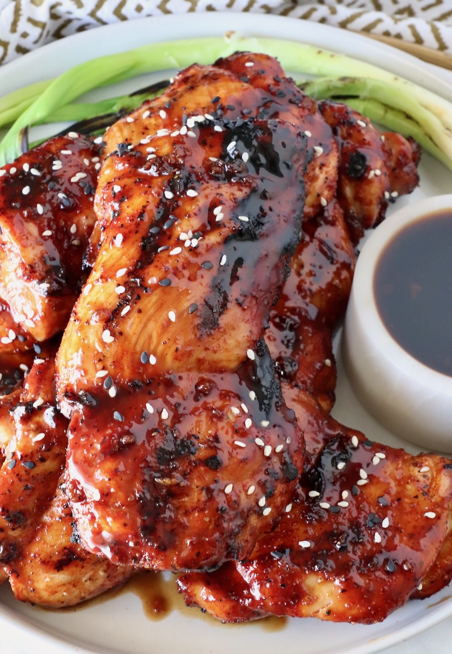 grilled chicken thighs stacked up on a plate glazed with bbq sauce, with green onions and Korean bbq sauce on the side of the plate