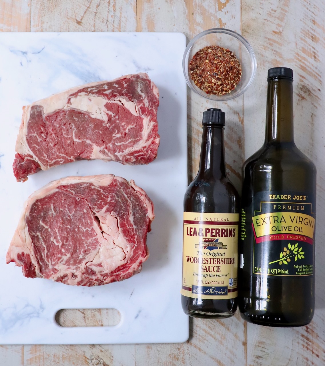 raw ribeye steaks on a cutting board, next to a bottle of Worcestershire sauce, a bottle of olive oil, and steak seasoning in a small glass bowl