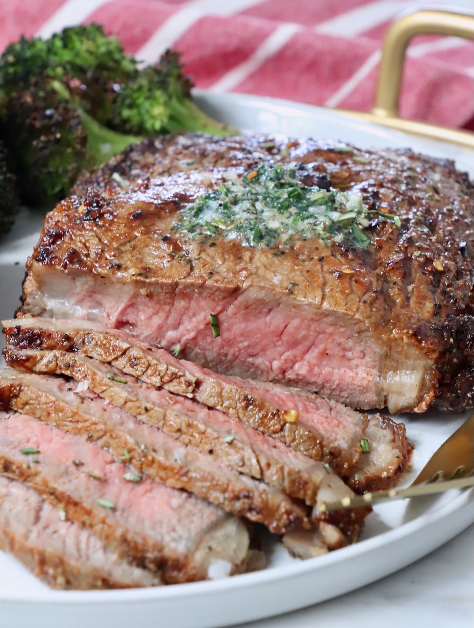 sliced ribeye steak on a plate with herb butter on top of the steak and roasted broccoli in the background on the plate