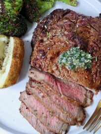 partially sliced ribeye steak on a plate with herb butter on top, cooked broccoli and a baked potato on the side