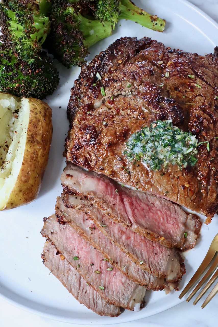 partially sliced ribeye steak on a plate with herb butter on top, cooked broccoli and a baked potato on the side
