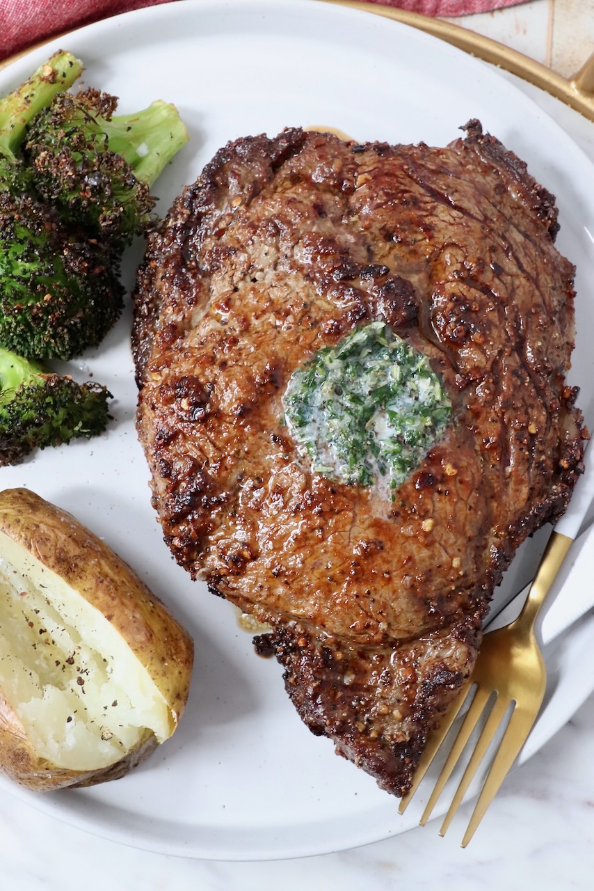 cooked ribeye steak on a plate with a fork, baked potato and cooked broccoli