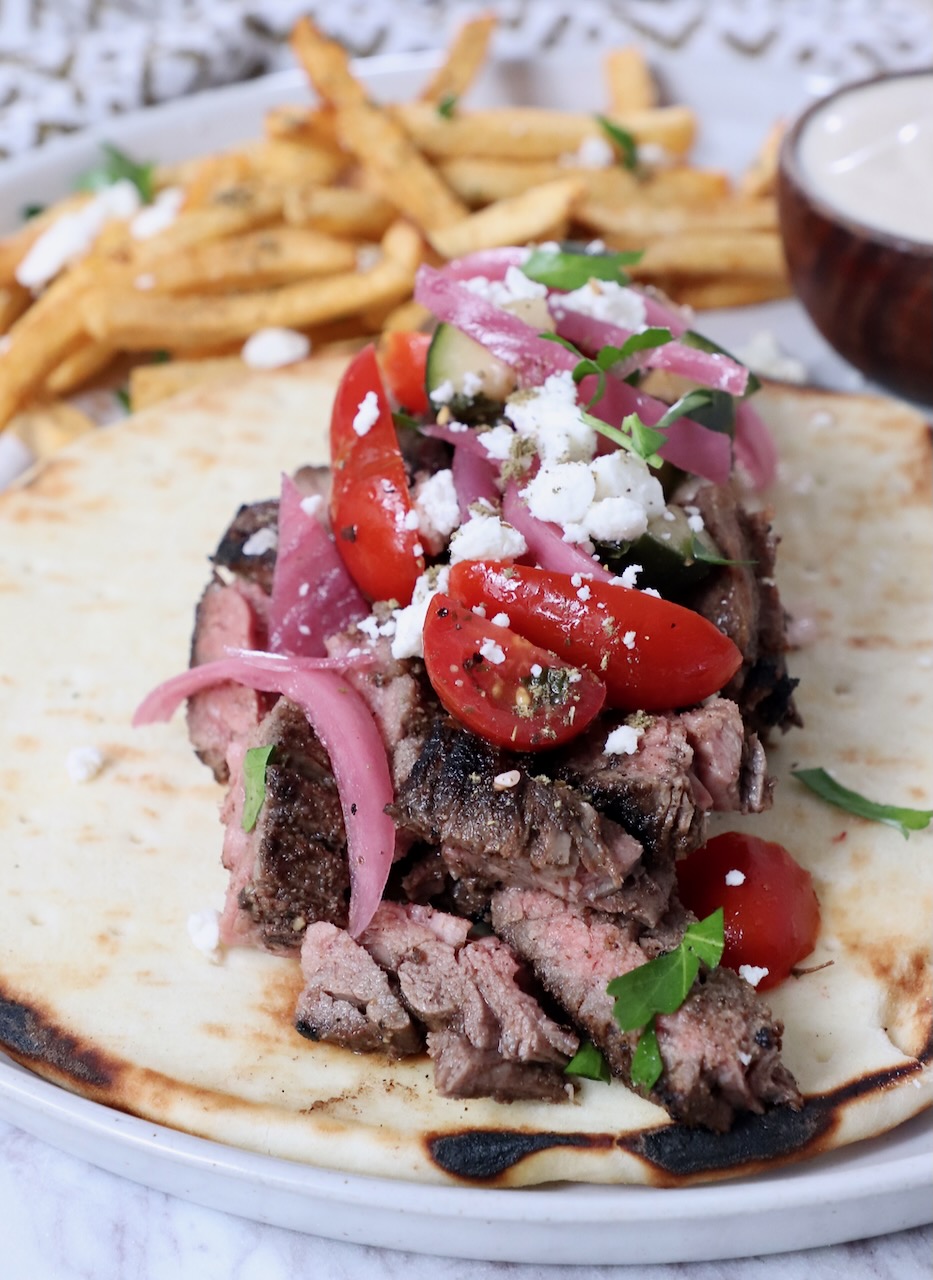 sliced, grilled steak on top of pita bread, topped with tomatoes, onions and feta cheese, on a plate with french fries