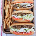 three chopped cheese sandwiches with sliced tomatoes and shredded lettuce lined up on a baking sheet with seasoned, cooked french fries on the side