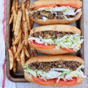 three chopped cheese sandwiches with sliced tomatoes and shredded lettuce lined up on a baking sheet with seasoned, cooked french fries on the side