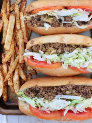 three chopped cheese sandwiches with sliced tomatoes and shredded lettuce lined up on a baking sheet with seasoned, cooked french fries on the side