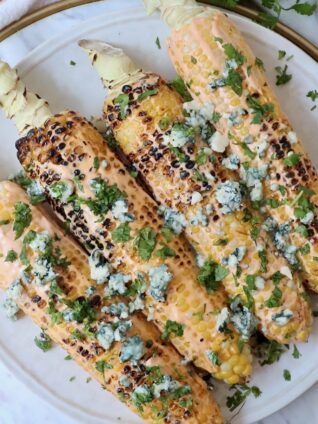 grilled corn on the cob on a plate topped with blue cheese crumbles and fresh chopped cilantro