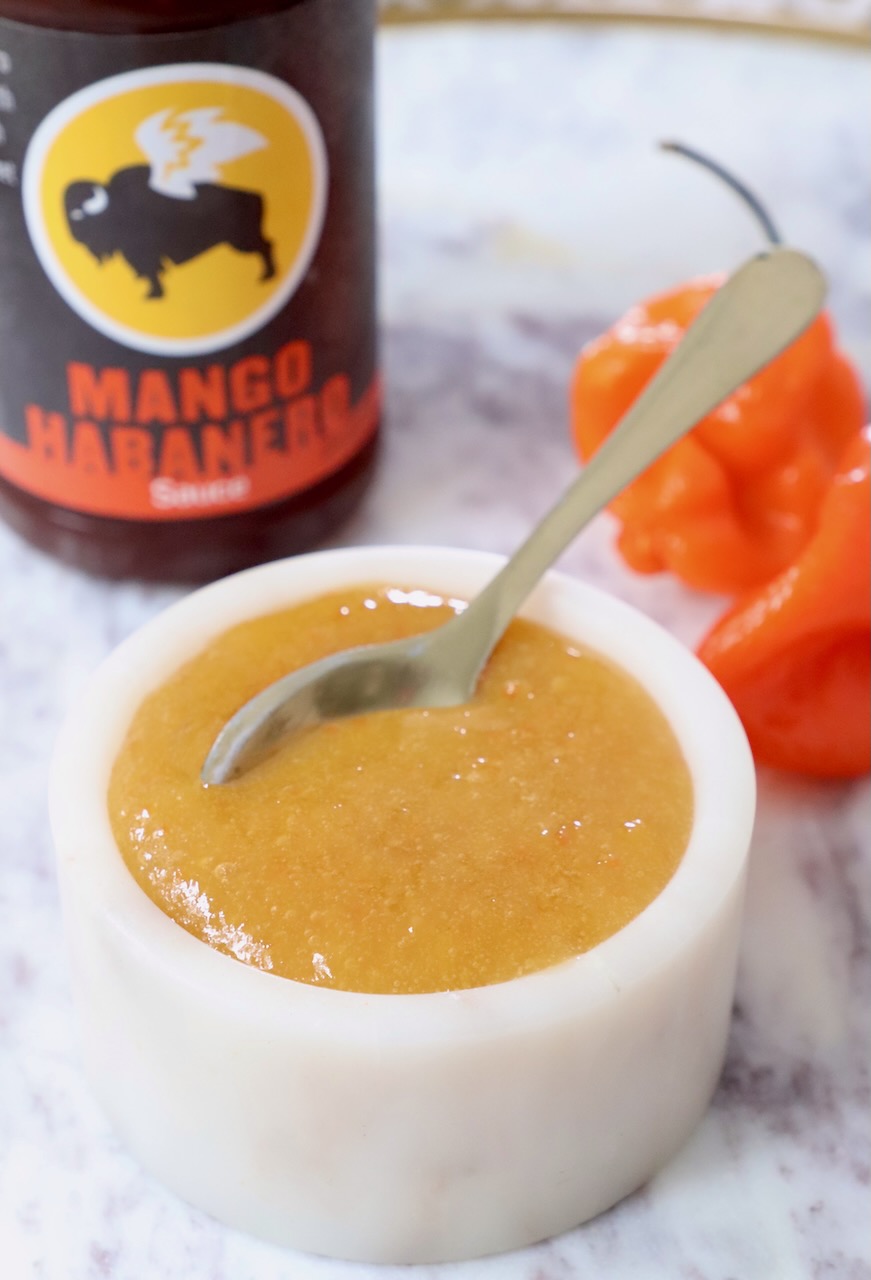 mango habanero sauce in a bowl with a spoon, with two habaneros behind it and a bottle of buffalo wild wings sauce