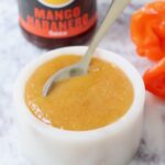 mango habanero sauce in a bowl with a spoon with a bottle of sauce in the background and fresh habanero peppers