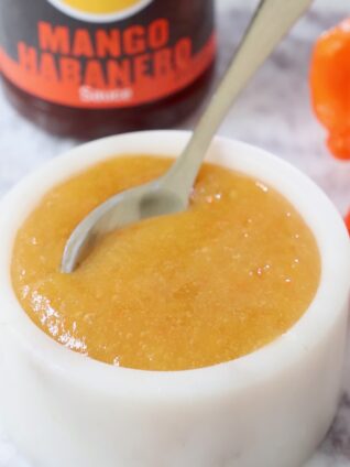 mango habanero sauce in a bowl with a spoon with a bottle of sauce in the background and fresh habanero peppers