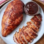 whole smoked chicken breast on a plate next to a sliced smoked chicken breast and a small bowl of bbq sauce