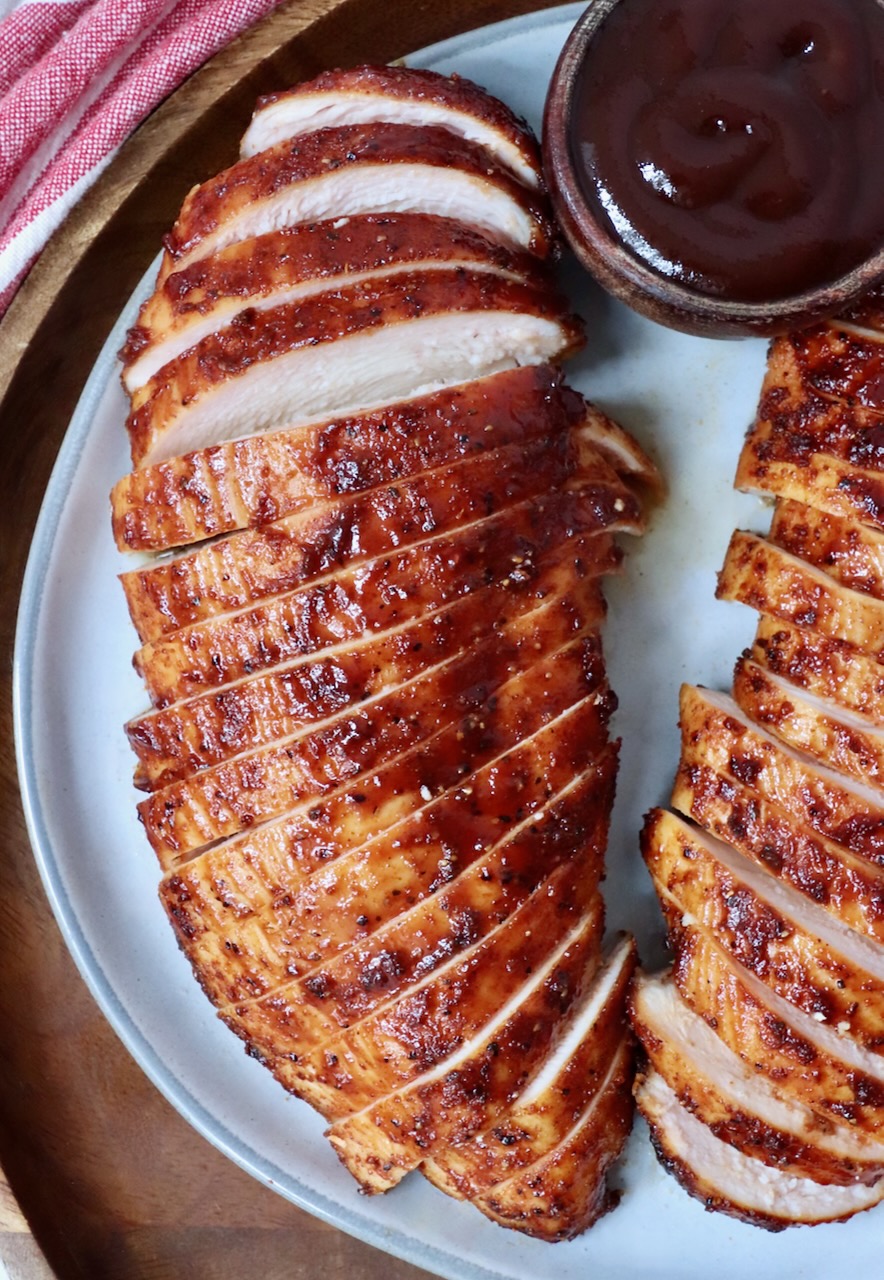 sliced smoked chicken breasts on a plate with a small bowl of bbq sauce on the side