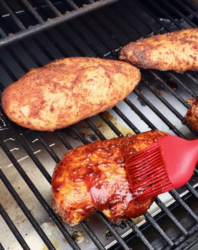 red brush with bbq sauce being brushed on a chicken breast on a smoker