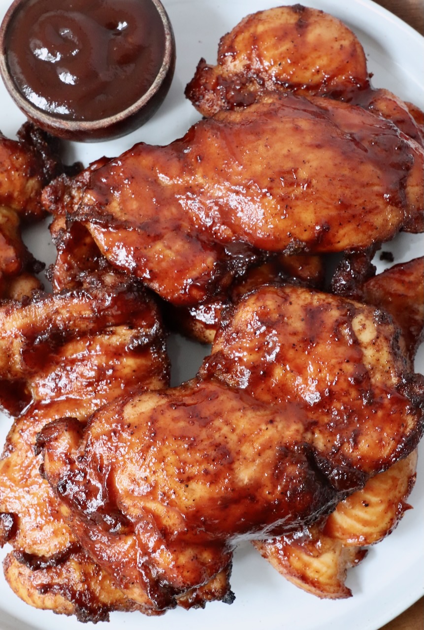 smoked chicken thighs glazed with bbq sauce stacked up on a plate with a small bowl of bbq sauce on the side