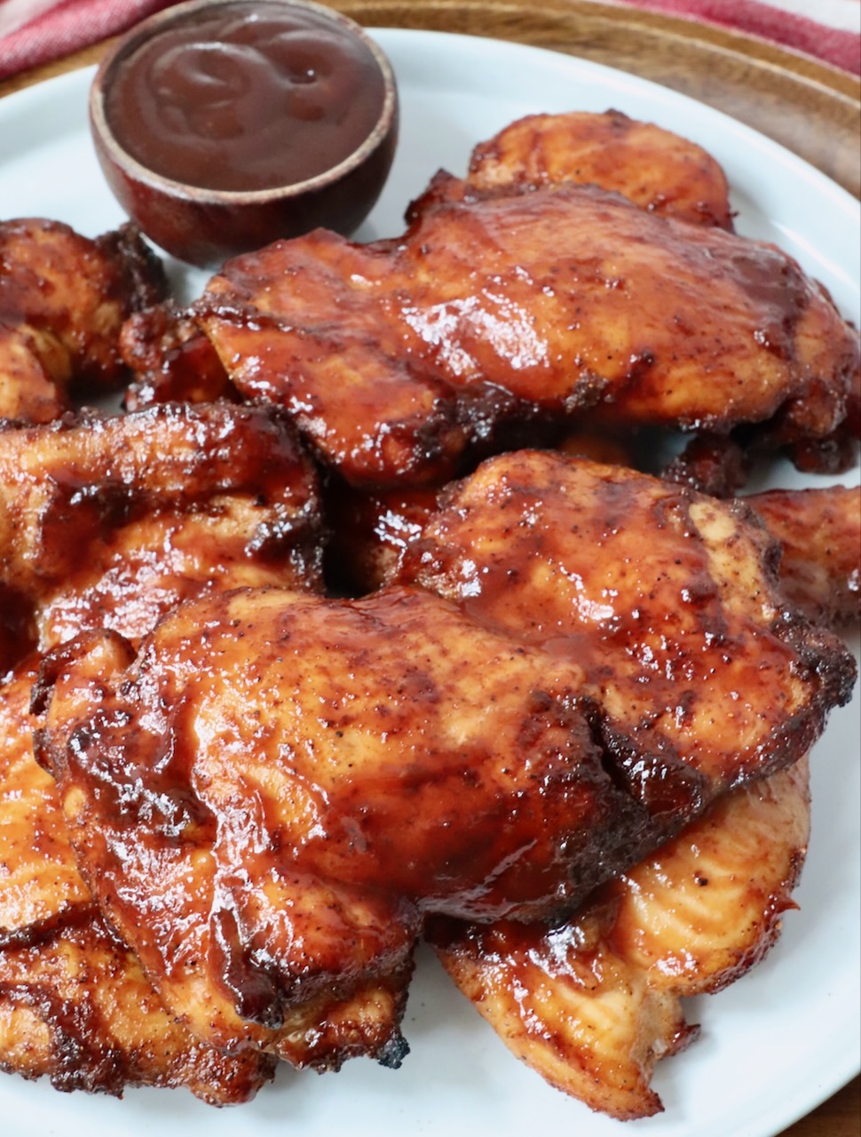 smoked chicken thighs stacked up on a plate with a small bowl of bbq sauce on the side