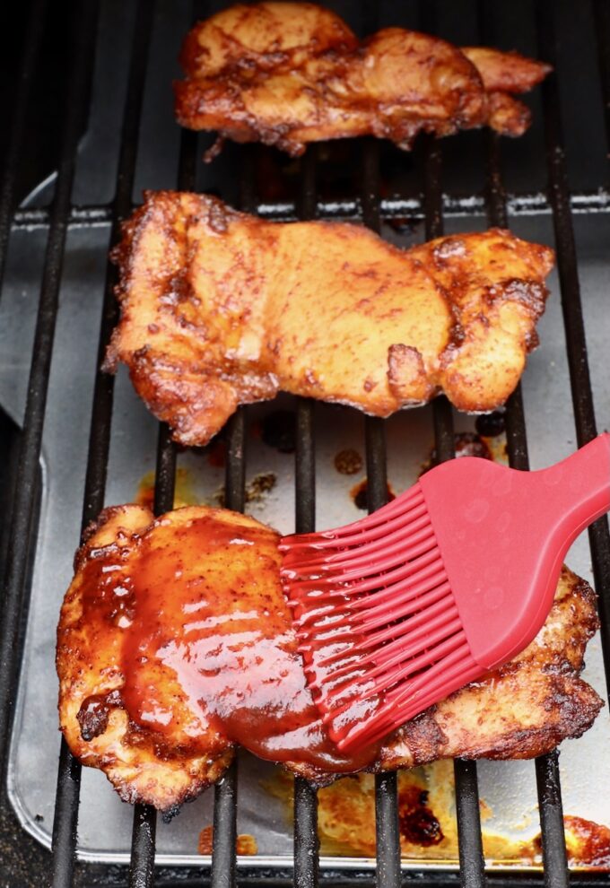 chicken thighs brushed with bbq sauce with a red brush on a smoker