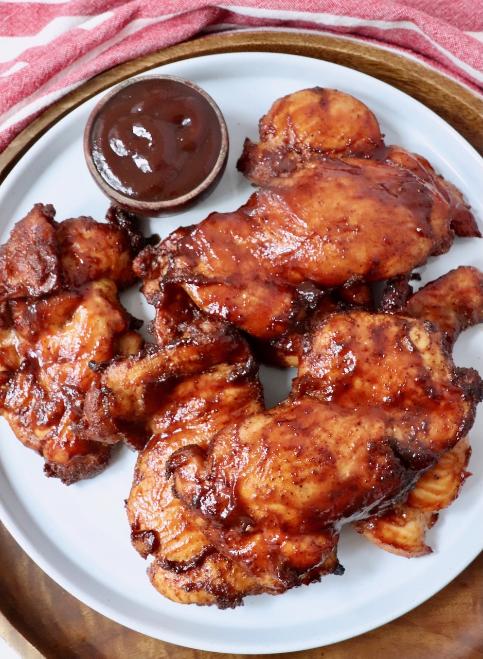 smoked chicken thighs stacked up on a plate with a small bowl of bbq sauce on the side