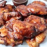 smoked chicken thighs stacked up on a plate with bbq sauce in a small bowl on the side