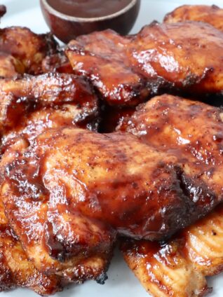 smoked chicken thighs stacked up on a plate with bbq sauce in a small bowl on the side