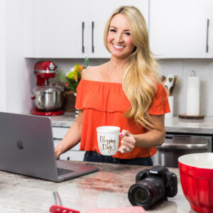 woman holding a mug in a kitchen standing next to an open laptop and canon camera