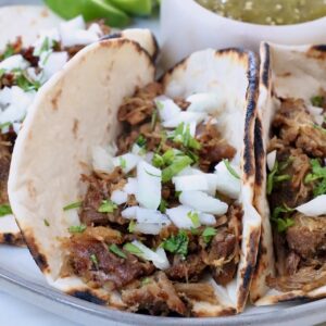 shredded pork in tortillas on plate, topped with diced onions and fresh chopped cilantro