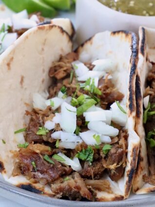 shredded pork in tortillas on plate, topped with diced onions and fresh chopped cilantro