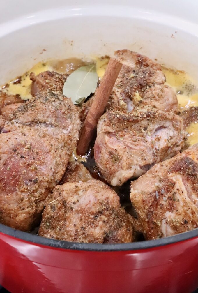 seasoned pieces of pork in a large cast iron dutch oven with liquid, cinnamon sticks and bay leaves