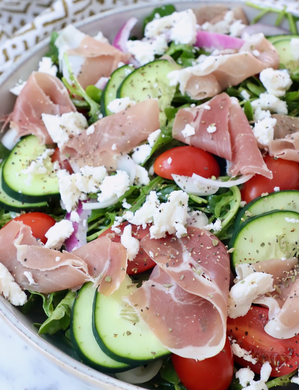 prosciutto, cucumber slices and tomatoes on top of a salad in a bowl