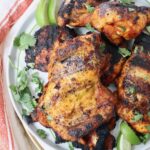 grilled chicken thighs stacked up on a plate with fresh cilantro and lime wedges