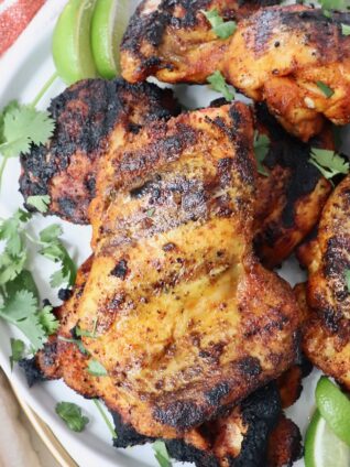 grilled chicken thighs stacked up on a plate with fresh cilantro and lime wedges