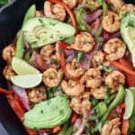 shrimp fajitas in a cast iron skillet topped with sliced avocado and lime wedges