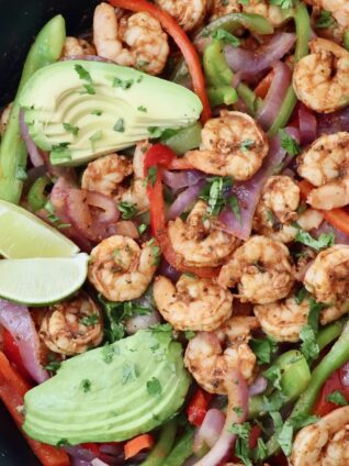 shrimp fajitas in a cast iron skillet topped with sliced avocado and lime wedges