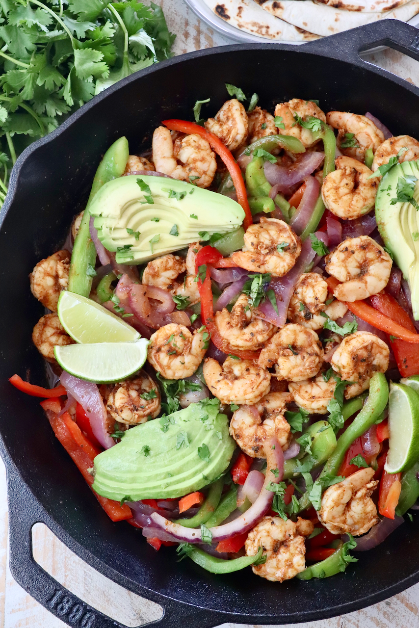 shrimp fajitas in a cast iron skillet topped with sliced avocado and lime wedges