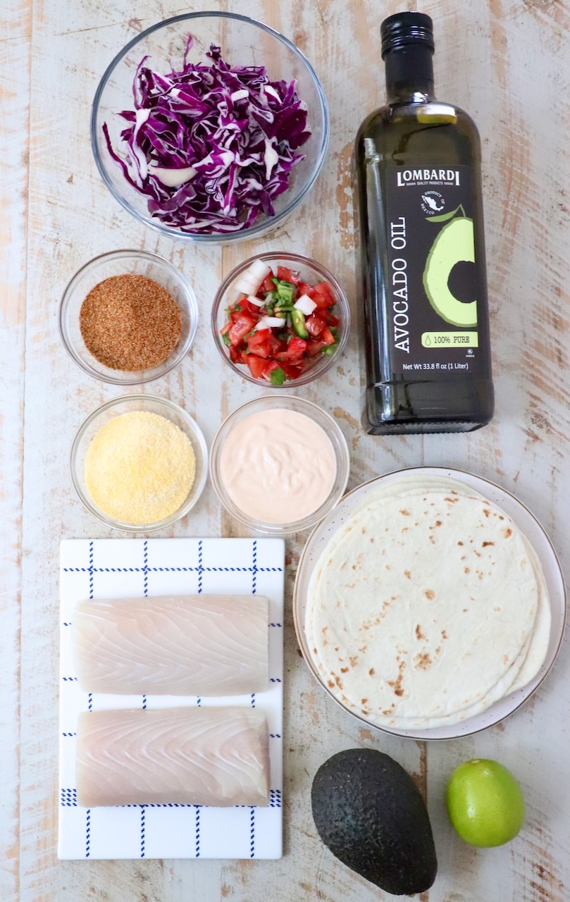 Top-down view of fish taco ingredients on a light wooden surface: red cabbage, avocado oil, taco seasoning, pico de gallo, fish taco sauce, cornmeal, tortillas, fish fillets, avocado, and a lime.