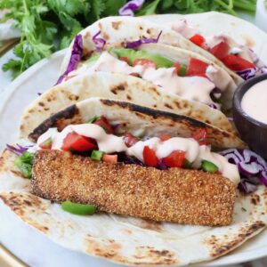 Three tacos with crispy fish, purple cabbage, diced tomatoes, and creamy sauce on a tortilla. With fresh cilantro in the background