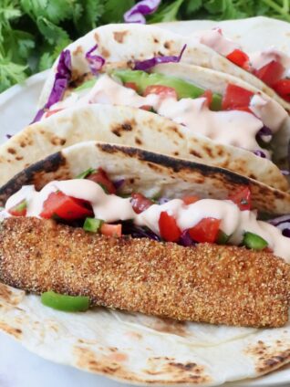 Three tacos with crispy fish, purple cabbage, diced tomatoes, and creamy sauce on a tortilla. With fresh cilantro in the background