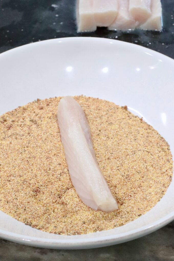 A raw fish fillet sits on seasoned cornmeal in a white bowl, ready for breading. In the background, additional fish fillets are visible on a dark surface.