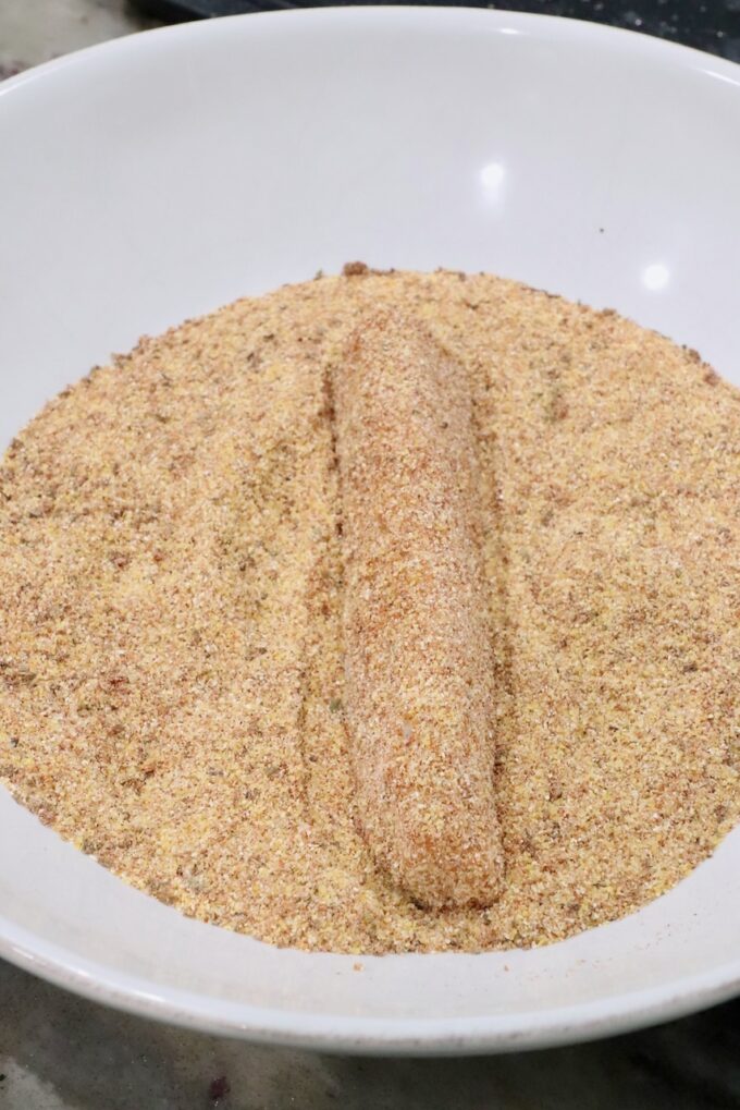 A breaded strip of fish lies on a bed of seasoned cornmeal in a white bowl.