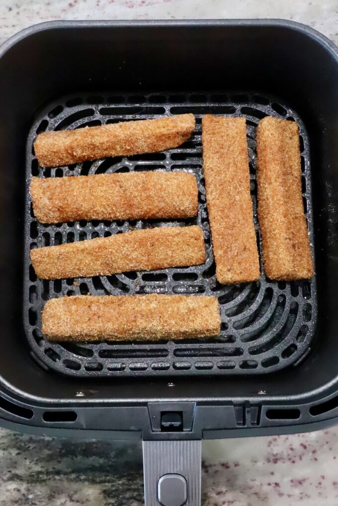 Six breaded fish sticks arranged in an air fryer basket on a kitchen counter. 