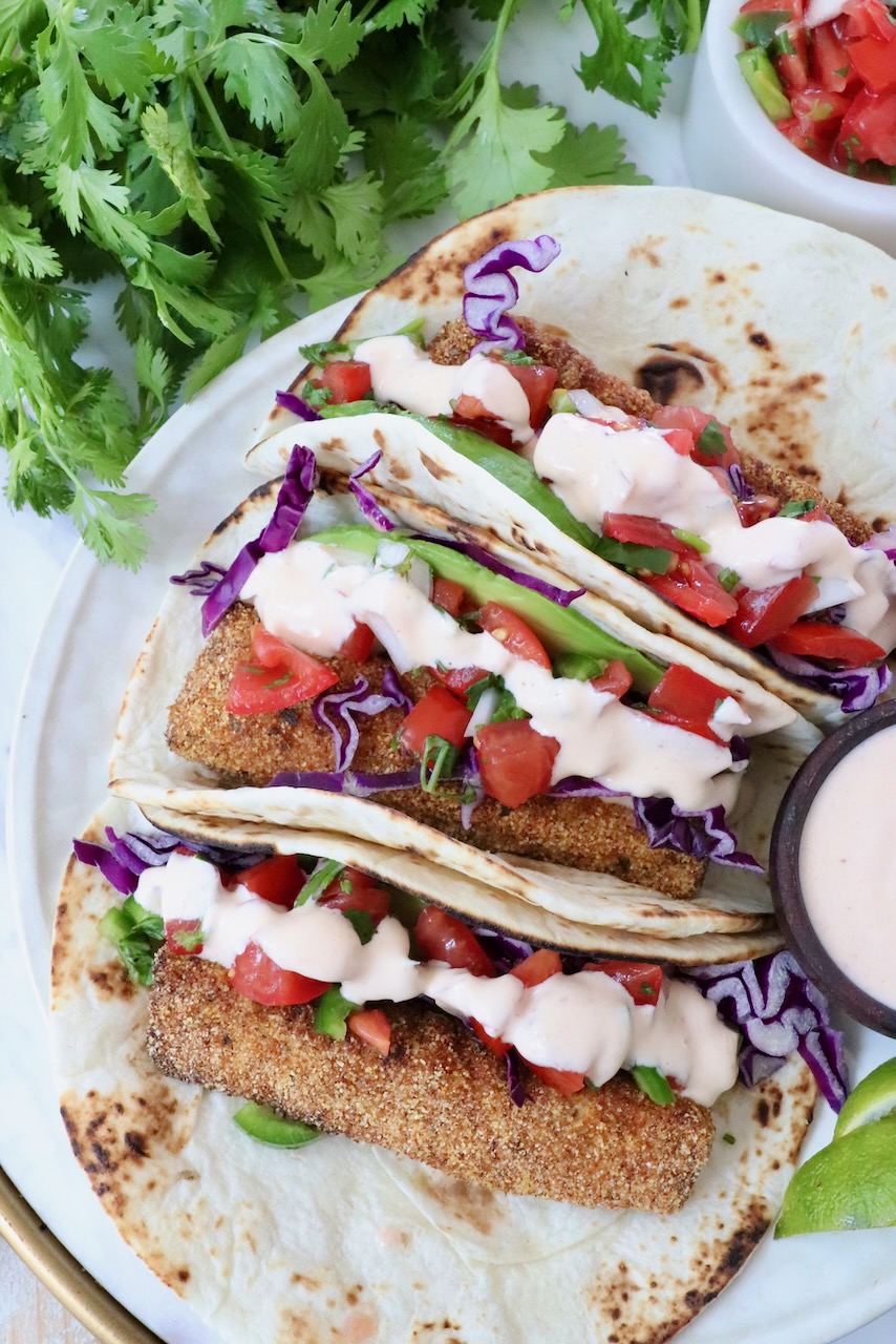 Three tacos with crispy fish, purple cabbage, diced tomatoes, and creamy sauce on a tortilla. They're garnished with fresh cilantro. 