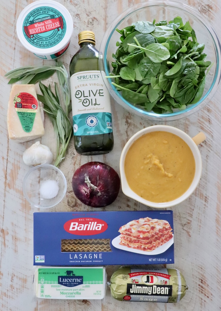 Ingredients for a lasagna dish arranged on a wooden surface. Items include ricotta cheese, olive oil, fresh spinach, mozzarella, lasagna noodles, and sausage.