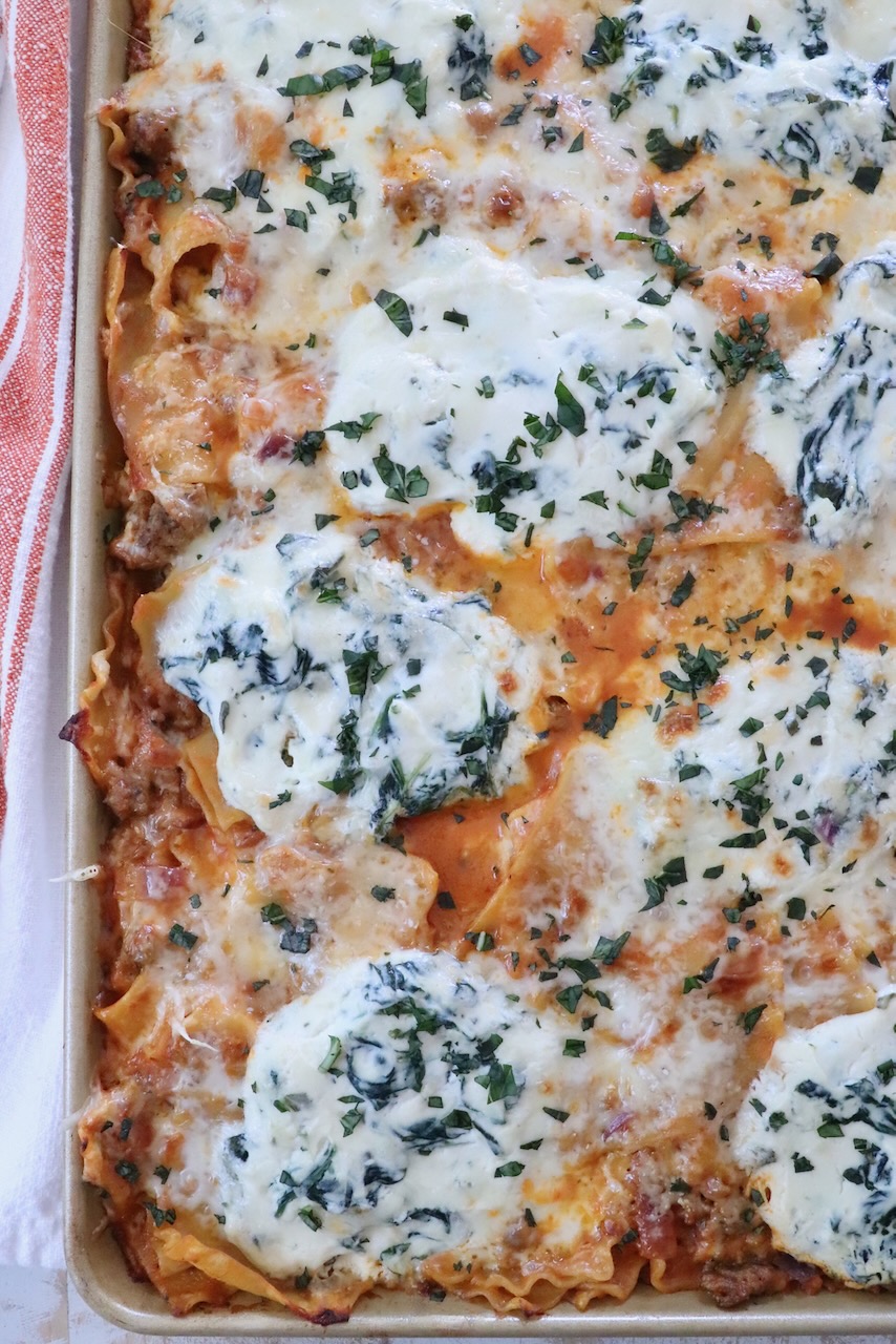 Close-up of a freshly-baked lasagna in a tray, topped with melted cheese, spinach, herbs, and a golden crust