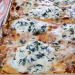 Close-up of a baked pasta dish in a rectangular tray, topped with bubbling cheese and herbs.
