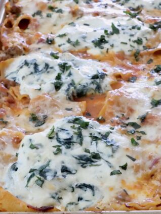Close-up of a baked pasta dish in a rectangular tray, topped with bubbling cheese and herbs.