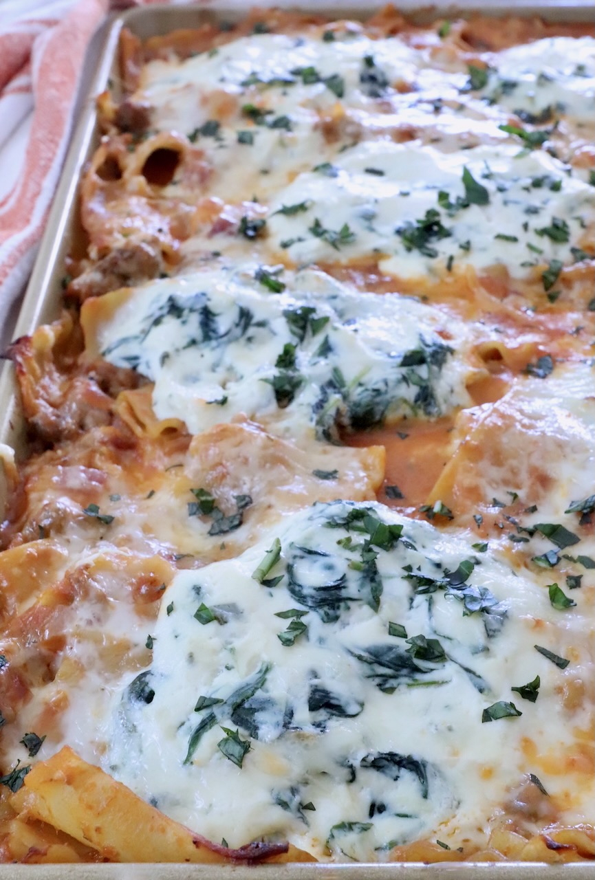 Close-up of a baked pasta dish in a rectangular tray, topped with bubbling cheese and herbs.
